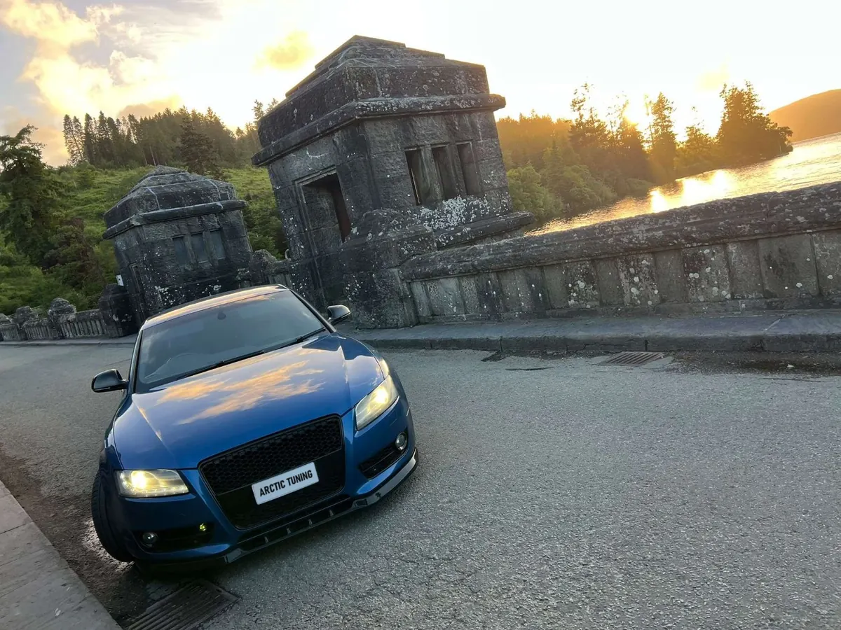 A blue Audi A5 with its headlights on, parked on a stone dam at sunset