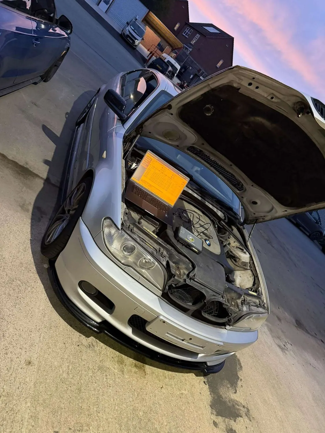A silver BMW coupé at dusk with its bonnet up and a laptop and remap tool working on the engine