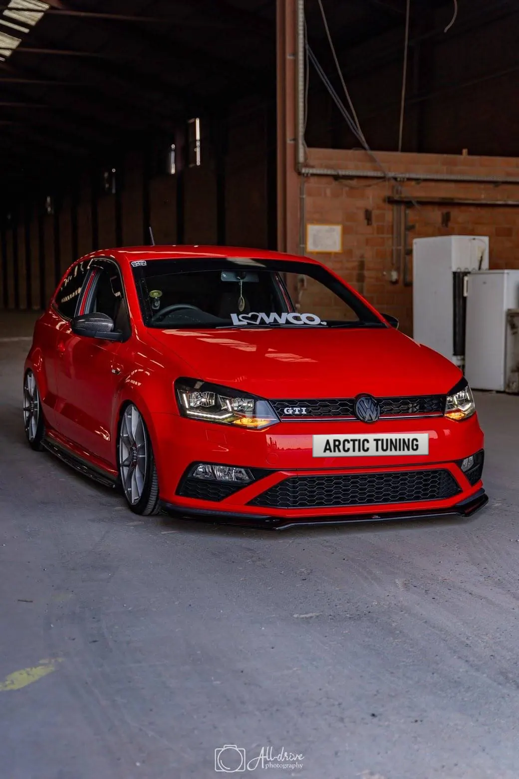 A lowered red Volkswagen Polo GTI with its lights on in an empty warehouse